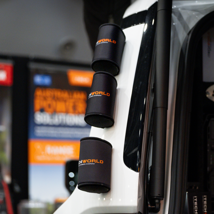 Three black iTechworld Magnetic Stubby Holders (375mL) with orange 4X4 WORLD logos are magnetically attached vertically to a white vehicle. A blurred indoor display with orange and blue signage is in the background.