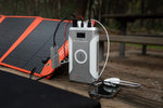 A Medicharge Flex | CPAP Battery | 276Wh sits on a wooden picnic table, powering a CPAP machine, smartphone, and solar panel—an ideal portable power supply for off-grid outdoor adventures.