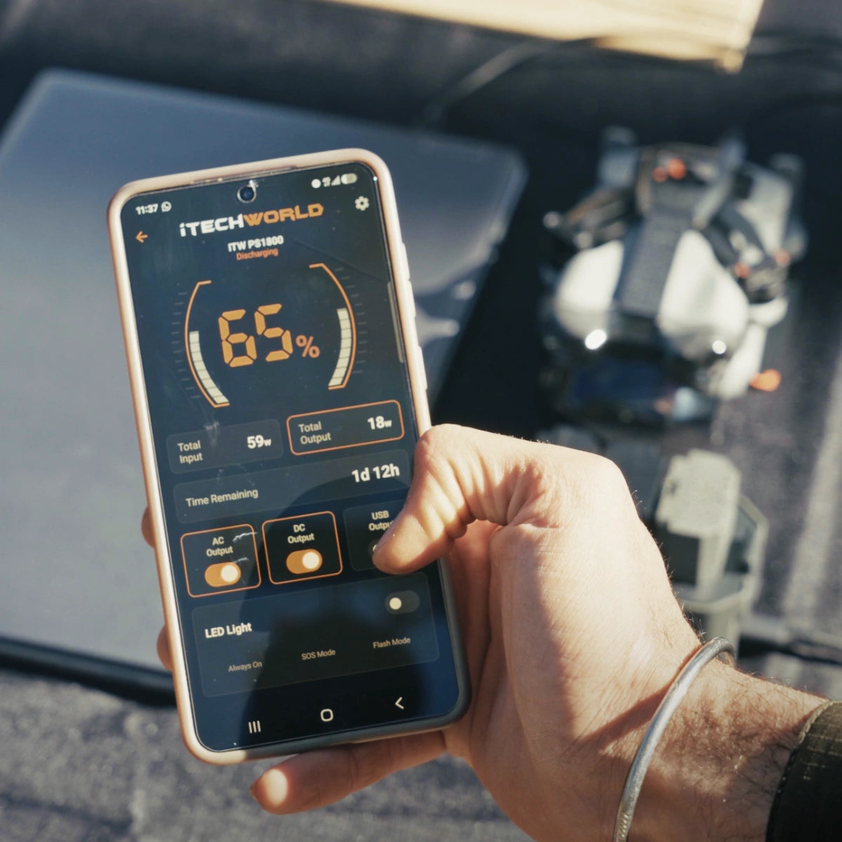 A hand holds a smartphone displaying a power management app with 65% battery, while an iTechworld Ultimate | Solar Generator Kit PS1800 PRO Power Station and 300W Solar Blanket, along with a drone and other electronic devices, appear blurred in the background.