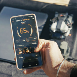 A hand holds a smartphone displaying a power management app with 65% battery, while an iTechworld Ultimate | Solar Generator Kit PS1800 PRO Power Station and 300W Solar Blanket, along with a drone and other electronic devices, appear blurred in the background.