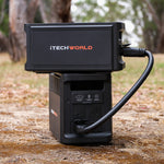 An iTechworld PS1800 PRO Portable Lithium Power Station (120Ah) in black sits outside on brown ground, plugged in with a thick cable, while blurred trees and greenery appear in the background.
