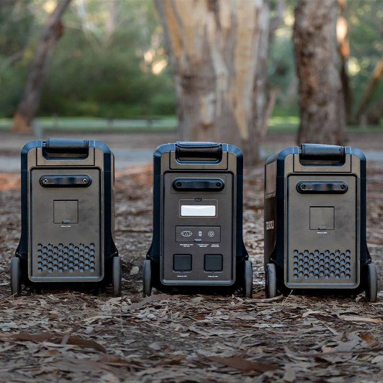 Three iTechworld PS3600 portable lithium power stations with EB3840 Expansion Battery Kits are lined up outdoors among trees and leaves, with the central unit displaying its control panels and ports.