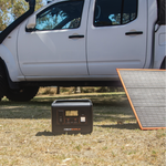 An iTechworld 800W 40Ah PS800 Portable Lithium Power Station and a solar panel are set up on the grass beside a white pickup truck outdoors, providing an ideal outdoor power supply. Trees and sunlight can be seen in the background.