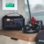 A lightweight iTechworld 300W 21.5Ah | PS300 Portable Lithium Power Station sits on a wooden surface beside black headphones, a red figurine, and a camera lens. Its compact design and 300W AC output make it ideal for powering your essentials. A green and white picture hangs on the wall above.