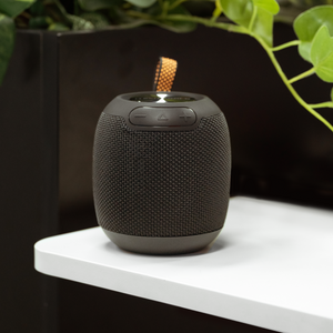 The iTechworld Pulse Mini Waterproof Portable Speaker, featuring textured sides and top control buttons, rests on a white surface near a black partition and green plants, delivering high-fidelity audio wherever you go.