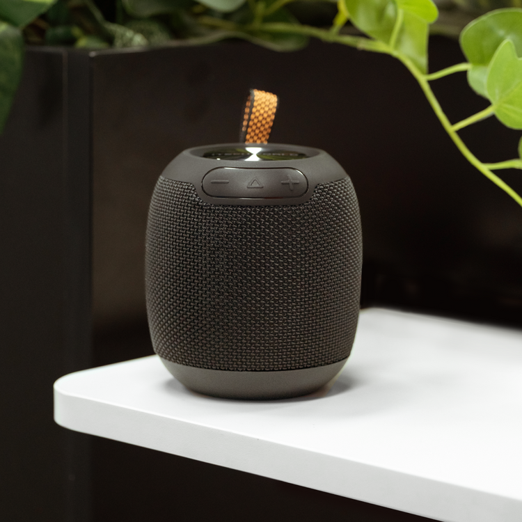 The iTechworld Pulse Mini Waterproof Portable Speaker, featuring textured sides and top control buttons, rests on a white surface near a black partition and green plants, delivering high-fidelity audio wherever you go.