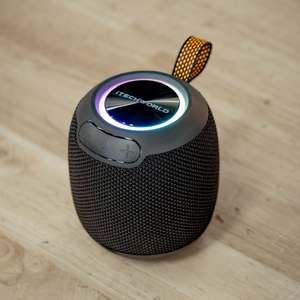 The iTechworld Pulse Mini is a small, black waterproof portable Bluetooth speaker with a mesh exterior, colorful LED light ring on top, control buttons, and an orange carrying loop, shown on a wooden surface.