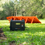 An iTechworld Elite PS2000 Power Station and 300W Solar Blanket sit on green grass in sunlight, with the orange foldable solar blanket behind the black power station and trees in the background.