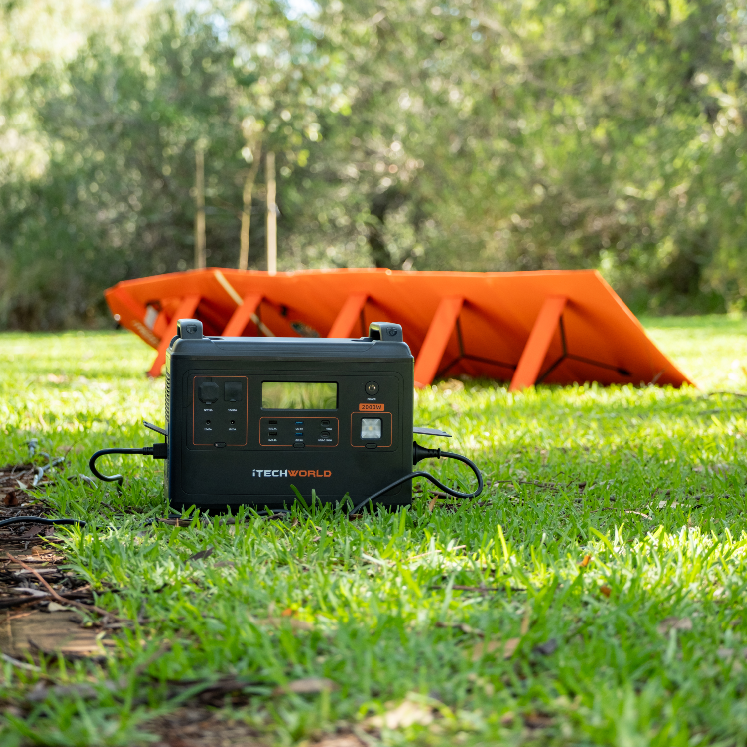 An iTechworld Elite Solar Generator Kit (PS2000 Power Station and 300W Solar Blanket) sits on grass, surrounded by trees and sunlight.