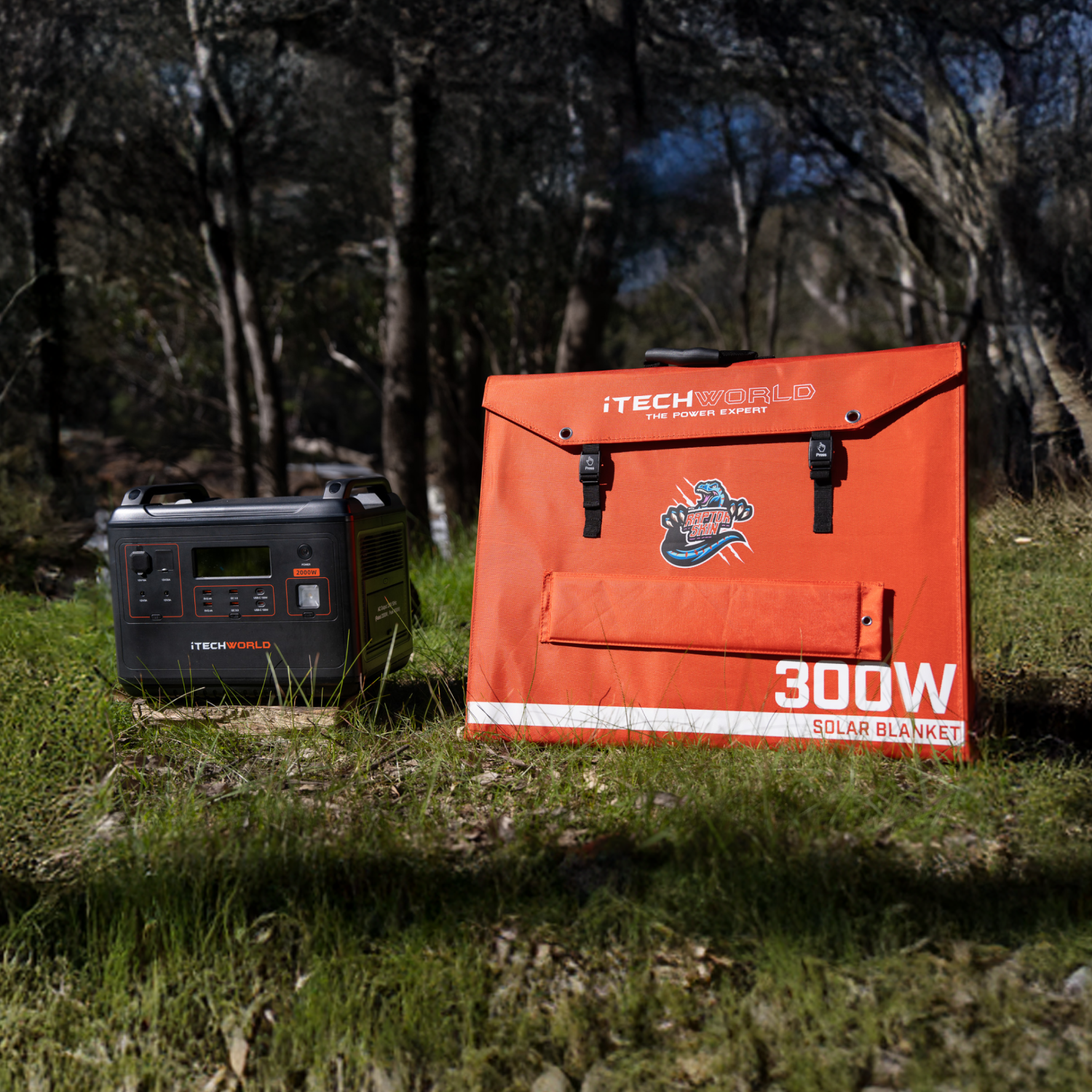 The iTechworld Elite Solar Generator Kit, featuring the PS2000 Power Station and an orange 300W Solar Blanket, is set on grass in a sunlit forest clearing with trees and dappled shadows in the background.