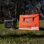 The iTechworld Elite Solar Generator Kit, featuring the PS2000 Power Station and a 300W Solar Blanket, sits on grass amid tall trees in a sunlit forest clearing.