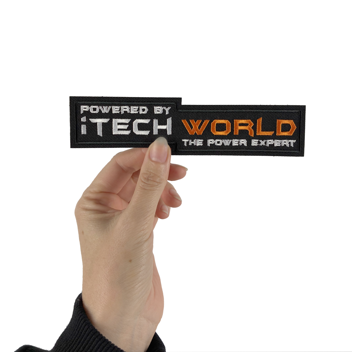 A hand holds the iTechworld Velcro Patch, featuring silver and orange “POWERED BY iTECHWORLD THE POWER EXPERT” text and the distinctive power expert logo on a black background.