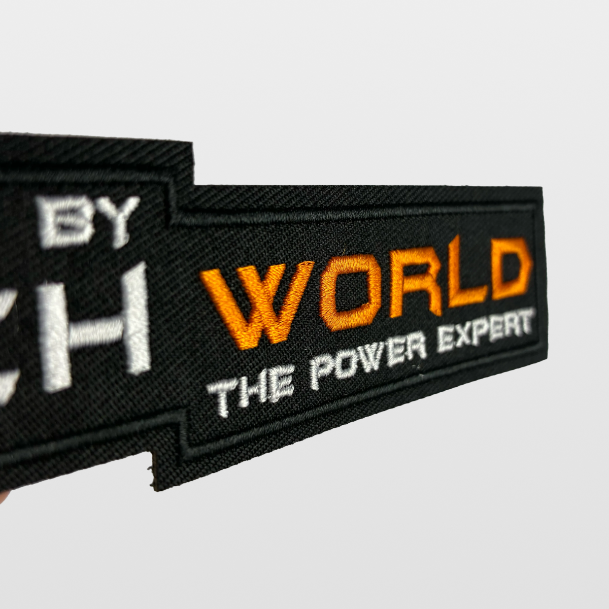 A close-up of the iTechworld Velcro Patch, featuring black fabric with “WORLD THE POWER EXPERT” embroidered in orange and white letters, displays the distinctive iTechworld logo against a plain light background.
