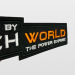 A close-up of the iTechworld Velcro Patch, featuring black fabric with “WORLD THE POWER EXPERT” embroidered in orange and white letters, displays the distinctive iTechworld logo against a plain light background.