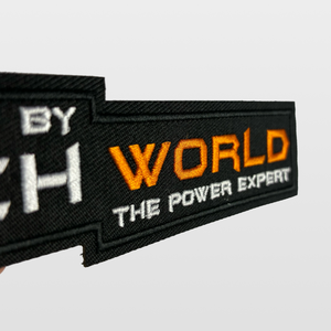A close-up of the iTechworld Velcro Patch, featuring black fabric with “WORLD THE POWER EXPERT” embroidered in orange and white letters, displays the distinctive iTechworld logo against a plain light background.
