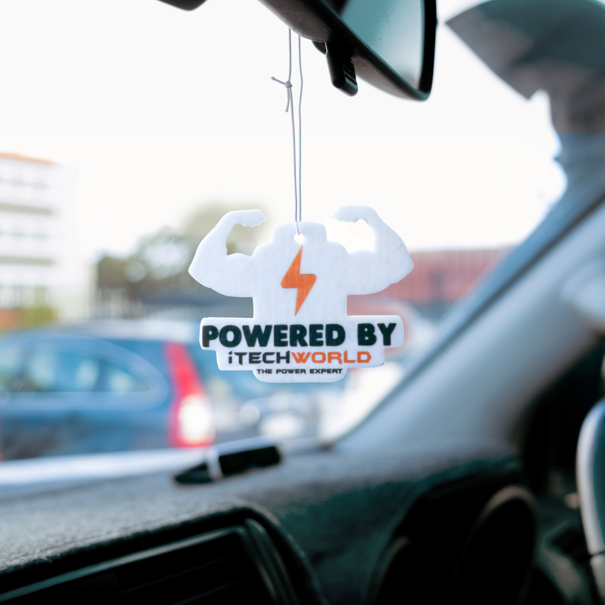 An iTechworld | Air Freshener | New Car Scent dangles from the rearview mirror with a flexed arm design and orange lightning bolt, displaying the iTechworld brand. Parked cars and buildings outside hint at its long-lasting fragrance.