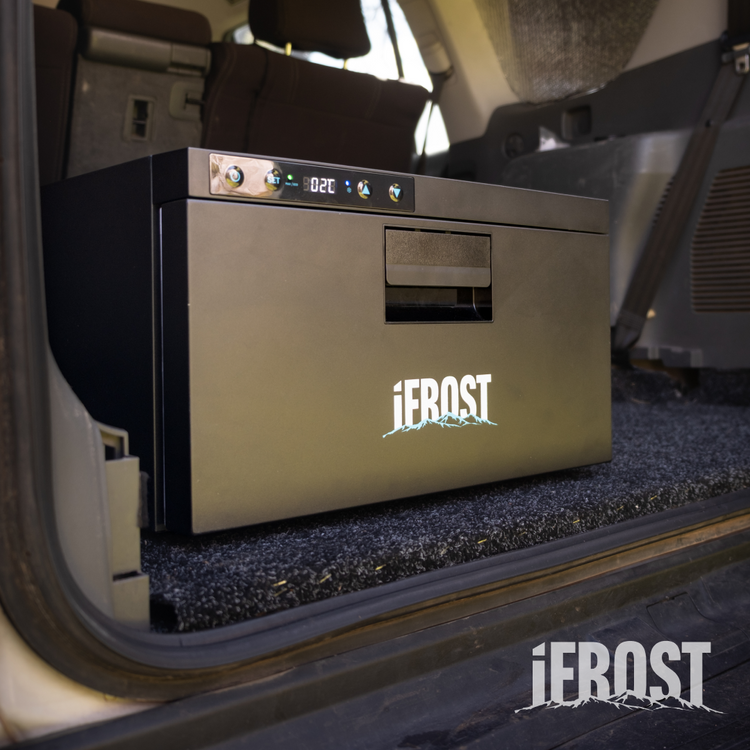 An iTechworld iFROST | Drawer Fridge Freezer | 31L with a digital display sits on a carpeted surface in the back of a vehicle, ready for travel or outdoor use. The iFROST logo is visible on the fridge and in the bottom right corner.
