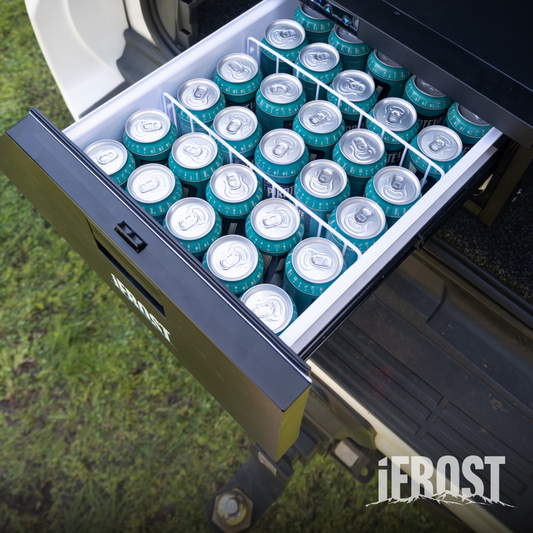 An open black iTechworld iFROST | Drawer Fridge Freezer | 31L, filled with neatly arranged drink cans, is attached to a 4WD over green grass. The fridge features an efficient LG Compressor for reliable cooling.