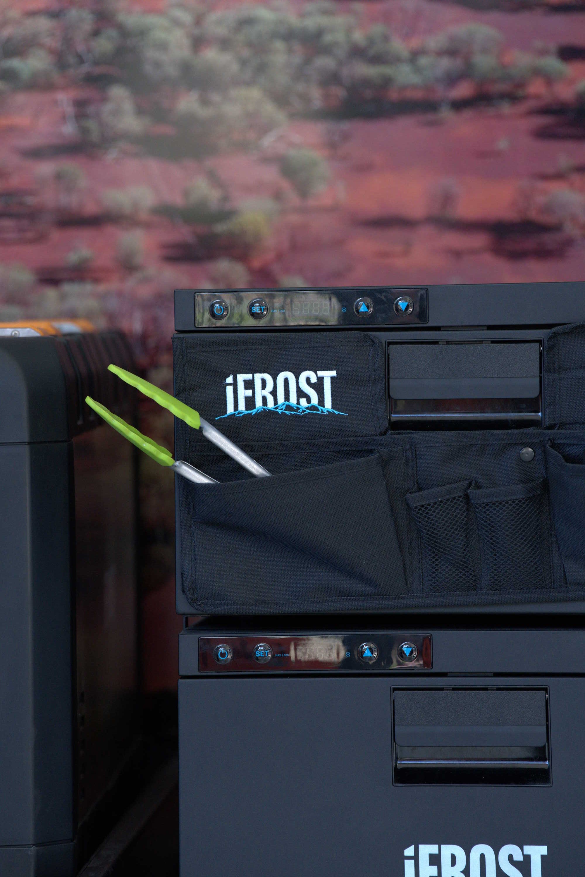 A close-up of two black iTechworld iFROST Draw Fridge Organisers, each featuring the iFROST label and a convenient pocket holding two pairs of green-handled tongs, serves as an ideal fridge storage solution set against a softly blurred nature-themed background.