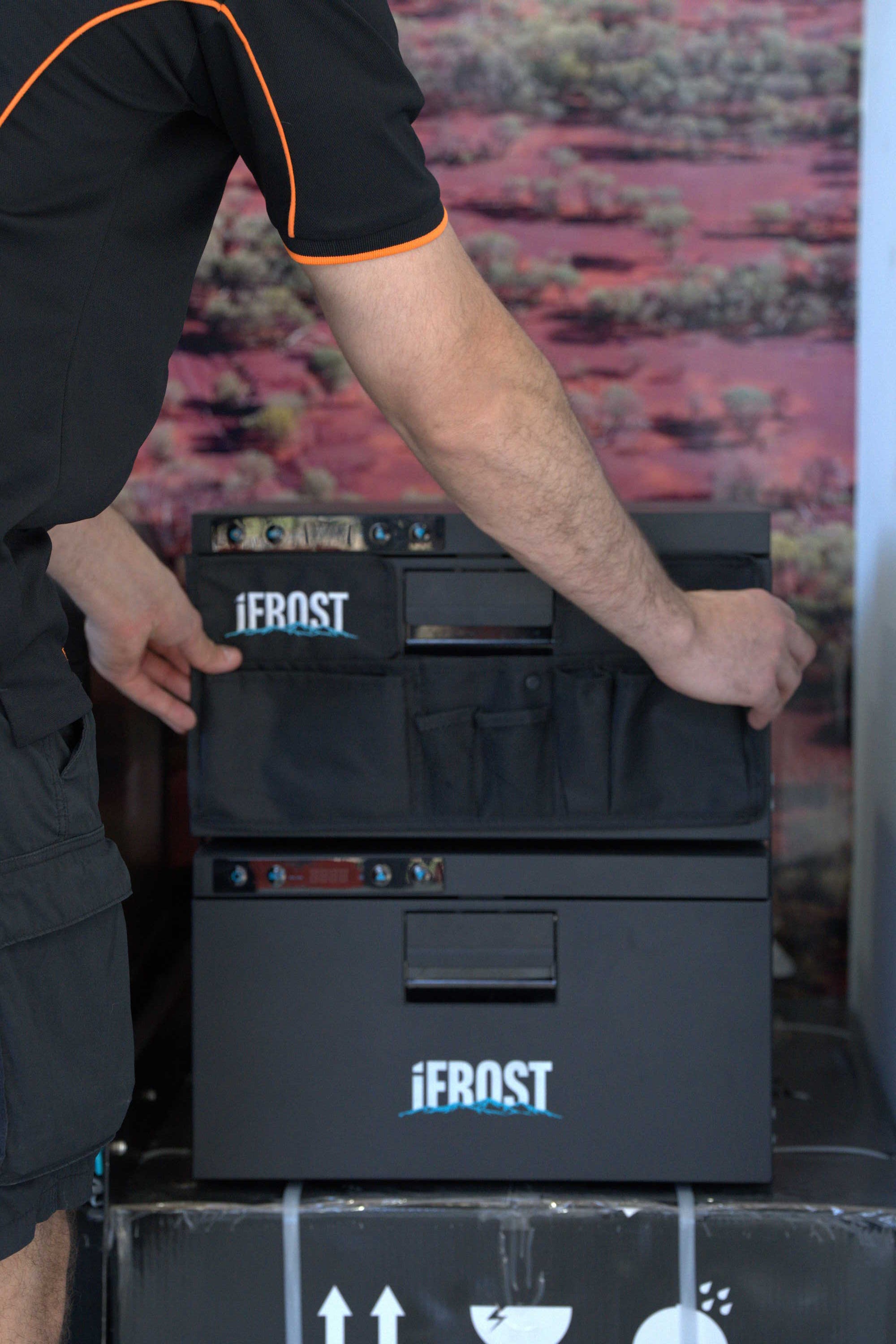 A person wearing a black shirt with orange trim is handling a black Jerost cooler or storage unit with multiple front compartments and visible Jerost branding.