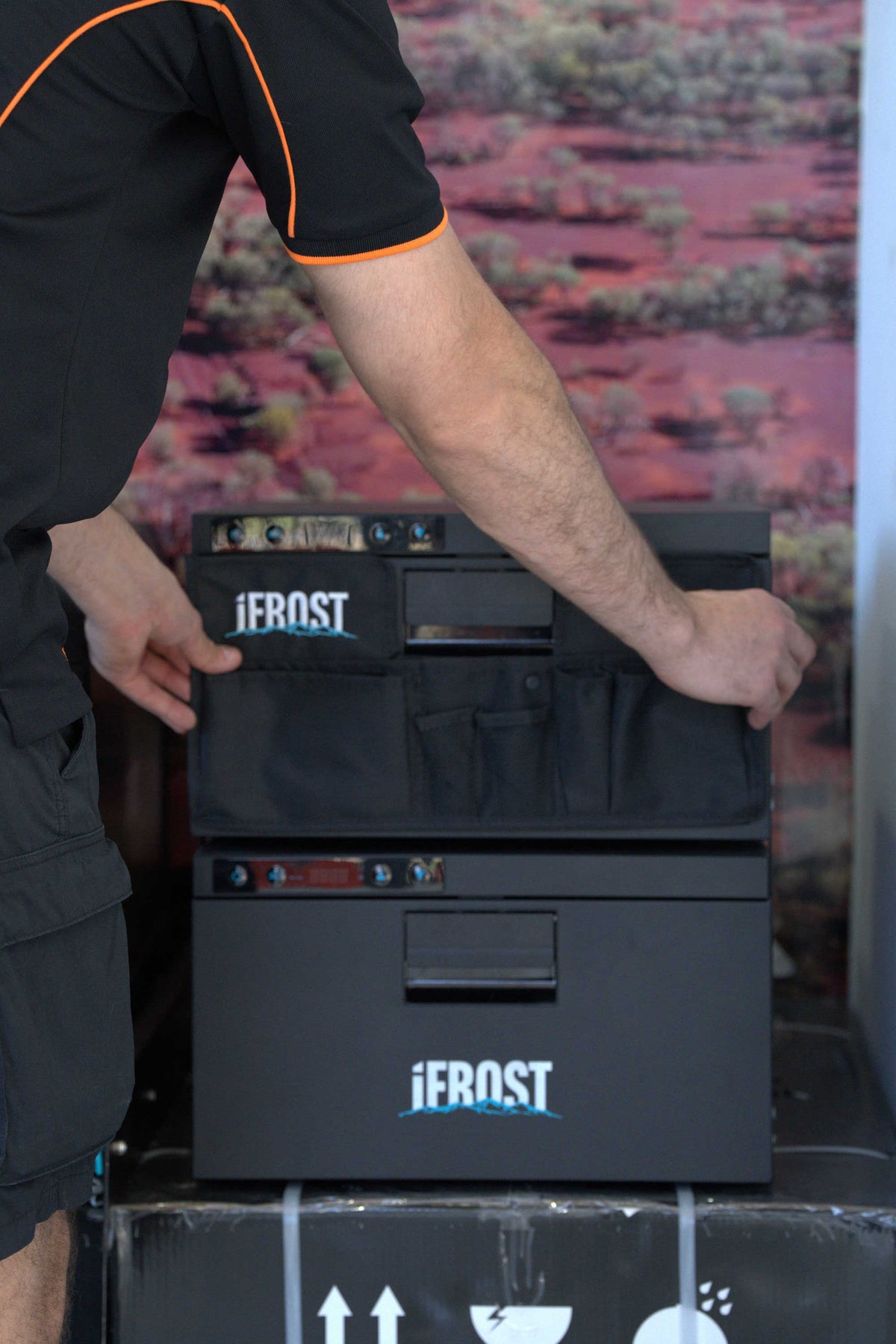 A person wearing a black shirt with orange trim is handling a black Jerost cooler or storage unit with multiple front compartments and visible Jerost branding.