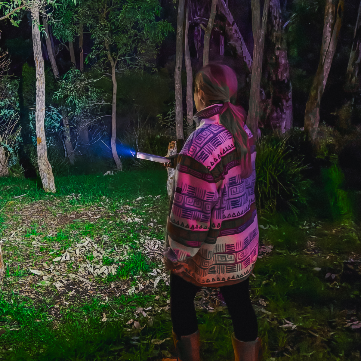 A person in a patterned jacket holds the iTechworld Mini Magnetic LED Strip Light (150LM), shining its bright beam into a forest at night, illuminating trees and the grassy, leaf-covered ground.