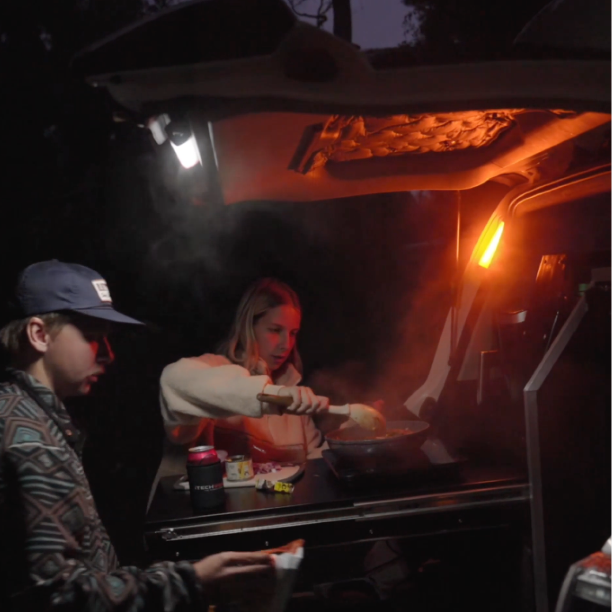A woman cooks on a stove in the back of a car at night, illuminated by orange and white light from the iTechworld Mini Magnetic LED Strip Light (150LM), while someone in a cap stands nearby holding an item. Steam rises from the pan.